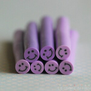 Puede incluir: Un conjunto de ocho cuentas de arcilla polimérica de color morado con una carita sonriente. Las cuentas son todas del mismo tamaño y forma, y tienen un simple diseño de carita sonriente negra.