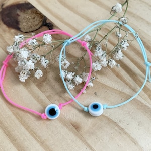 Peut inclure: Deux bracelets réglables, un rose et un bleu clair, chacun orné d'un charm en forme d'œil protecteur blanc et bleu. Les bracelets sont présentés sur une surface en bois clair avec de petites fleurs blanches.