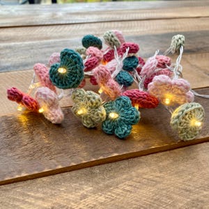 Pode incluir: Um fio de luzes LED com pequenas flores e corações feitos à mão em croché, em várias cores pastel, incluindo rosa claro, verde sálvia e azul-petróleo. Cada peça de croché envolve uma pequena lâmpada LED de luz branca quente.