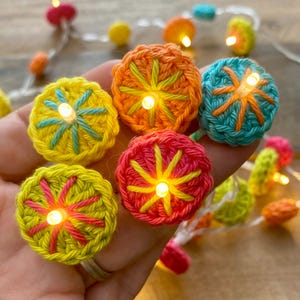 Op de afbeelding: Een lichtsnoer met kleine, ronde, handgehaakte hoesjes in levendige kleuren zoals geel, oranje, roze en turquoise. Elk gehaakt onderdeel heeft een stervormig geborduurd midden en wordt verlicht door een warm LED-lampje in het centrum.