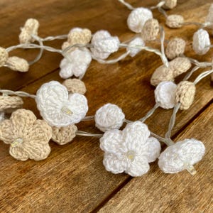 Pode incluir: Um fio de luzes LED com pequenas flores e corações de crochê em fios brancos e beges alternados. Cada peça de crochê está centrada em torno de uma pequena lâmpada LED transparente, criando um fio de iluminação decorativo.