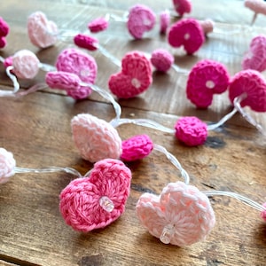 Può includere: Una stringa di luci a forma di cuore all'uncinetto in tonalità rosa e bianco. I cuori sono collegati da un filo trasparente e sono illuminati da piccole lampadine trasparenti. Le luci sono disposte su una superficie di legno.