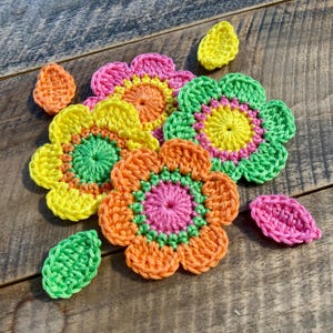 May include: A collection of colourful crochet flower appliqués and small leaves. The flowers feature layered petals in vibrant shades of pink, yellow, green, and orange. The individual leaves are crocheted in solid yellow, green, and pink yarn.
