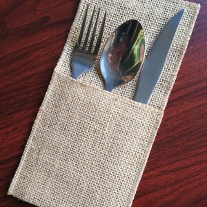 May include: A burlap cutlery holder containing a fork, spoon, and knife. The stainless steel utensils are arranged inside the beige fabric pouch. The holder is rectangular and rests on a dark wood surface.