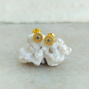 May include: A pair of gold stud earrings with a small blue gemstone in the centre of each earring.