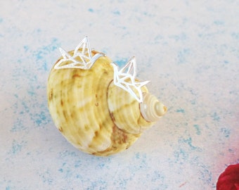 May include: Two silver-toned stud earrings shaped like geometric origami cranes. Each earring features a delicate, angular wireframe design that mimics the folded paper structure of a bird, resting on a tan and brown spiral seashell.