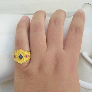 May include: A gold ring featuring a textured surface with a central square blue gemstone flanked by two smaller round pink gemstones. The band has an elongated hexagonal shape with intricate embossed patterns across the metal surface.