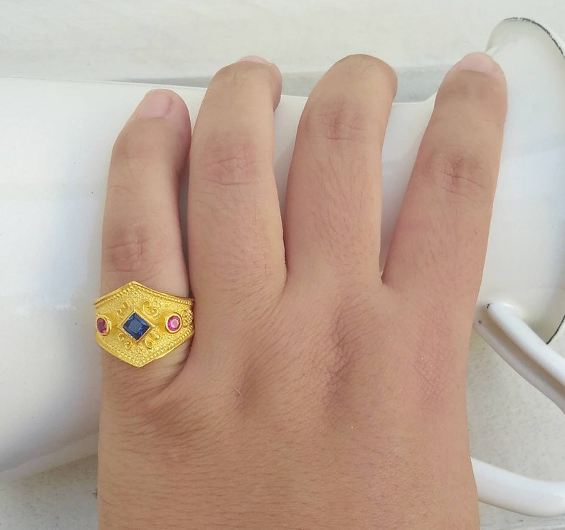 May include: A gold ring featuring a textured surface with a central square blue gemstone flanked by two smaller round pink gemstones. The band has an elongated hexagonal shape with intricate embossed patterns across the metal surface.