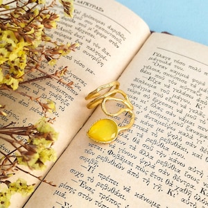 Könnte beinhalten: Ein goldfarbener Ring mit einer offenen, organisch strukturierten Ringschiene und einem einzelnen, facettierten, tropfenförmigen gelben Stein. Der Ring liegt auf einem aufgeschlagenen Buch mit griechischem Text neben gelben Trockenblumen.
