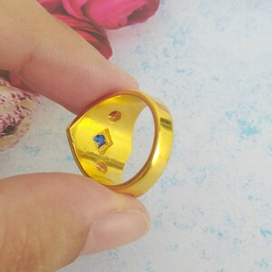 May include: A gold-toned ring featuring a smooth, polished band. The interior of the ring displays a small, square-cut blue gemstone set centrally, flanked by two smaller, circular, reddish-brown accents on either side of the blue stone.