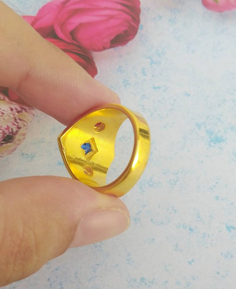 May include: A gold-toned ring featuring a smooth, polished band. The interior of the ring displays a small, square-cut blue gemstone set centrally, flanked by two smaller, circular, reddish-brown accents on either side of the blue stone.