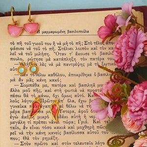 Puede incluir: Pendientes dorados con medias circunferencias de esmalte rosa y un par de pendientes rojos en forma de ala. Los pendientes se exhiben sobre un libro antiguo con flores rosas.
