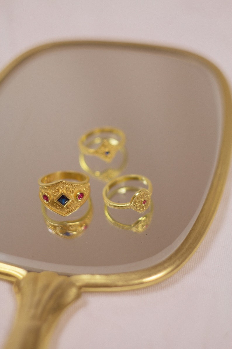 May include: Three gold-toned rings with textured surfaces. One ring features a central blue diamond-shaped stone flanked by two small red stones. Another ring has a circular face with a small red stone. The third ring is partially visible.