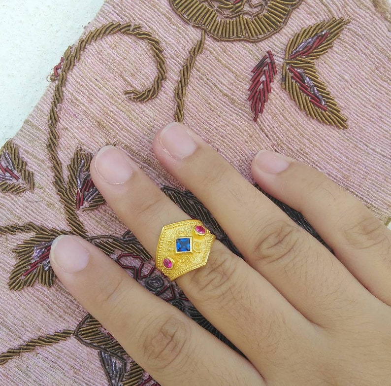May include: A gold ring featuring a central square blue gemstone flanked by two smaller oval pink gemstones. The band has a textured, matte gold finish with intricate embossed scrollwork patterns across the surface of the metal setting.