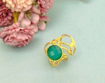 May include: A gold ring featuring a round, faceted green gemstone set in a textured, open-work band. The metal has a bright yellow finish, and the stone is a vibrant, translucent green with a geometric cut that reflects light across its surface.