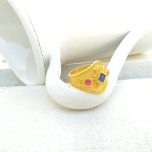 May include: A gold-toned ring featuring a textured, granulated surface with a central square-cut blue gemstone flanked by two smaller round-cut pink gemstones. The band has a pointed, shield-like front section with intricate metalwork details.