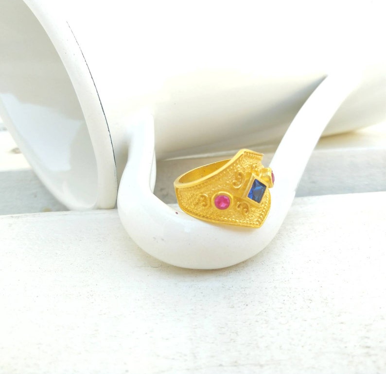 May include: A gold-toned ring featuring a textured, granulated surface with a central square-cut blue gemstone flanked by two smaller round-cut pink gemstones. The band has a pointed, shield-like front section with intricate metalwork details.