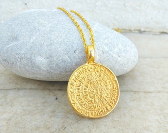 Peut inclure: Un pendentif circulaire doré en forme de disque de Phaistos, suspendu à une chaîne assortie. Le médaillon présente des symboles en spirale complexes et des inscriptions anciennes en relief gravées sur toute la surface métallique dorée.