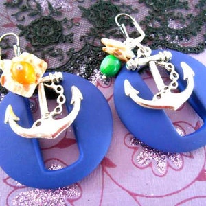 Könnte beinhalten: Ein Paar blaue und silberne Ohrringe mit Anker-Anhängern und einem kleinen orangefarbenen und grünen Perlenakzent. Die Ohrringe sind aus Polymer Clay gefertigt und haben einen einzigartigen, handgefertigten Look.