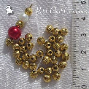 May include: A close-up of a pile of gold-colored metal beads with a ribbed texture. The beads are approximately 1 cm in diameter and are scattered on a white surface. A ruler is visible in the background.