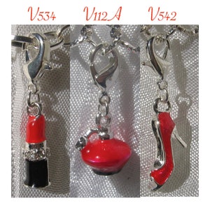 May include: Three silver-toned metal charms with lobster clasps, labeled V534, V112A, and V542. The charms feature a red and black lipstick tube with clear crystals, a red perfume bottle, and a red high-heeled shoe. Each charm hangs from a silver chain.