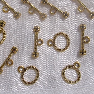 May include: A collection of gold-toned metal clasps and toggles for jewelry making. The clasps are round with a textured surface and have a small loop on one side. The toggles are straight with a textured surface and have a small loop on each end.