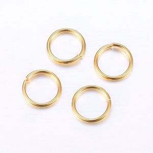 May include: Four small, gold-coloured split rings are arranged on a white surface. Each ring is circular with a small opening, allowing them to be opened and closed. The rings have a smooth, shiny finish, reflecting light.