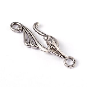 Puede incluir: Cierre de metal plateado con un dise&ntilde;o ornamentado. El cierre tiene un bucle en un extremo y un gancho en el otro.