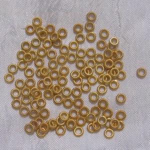 May include: A large quantity of small, gold-colored metal jump rings. The rings are scattered on a white fabric surface.