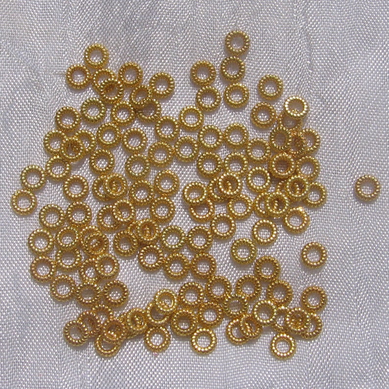 May include: A large quantity of small, gold-colored metal jump rings. The rings are scattered on a white fabric surface.