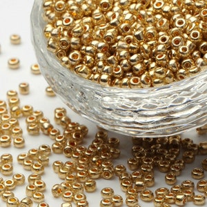May include: A bowl filled with gold-colored seed beads. The beads are scattered on a white surface.