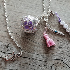 Puede incluir: Un collar de cadena plateada con un colgante de jaula que contiene un orbe de vidrio morado. El collar tambi&eacute;n tiene un borla rosa, un amuleto de pluma plateada y un amuleto con el nombre "LINA" escrito en cuentas blancas.