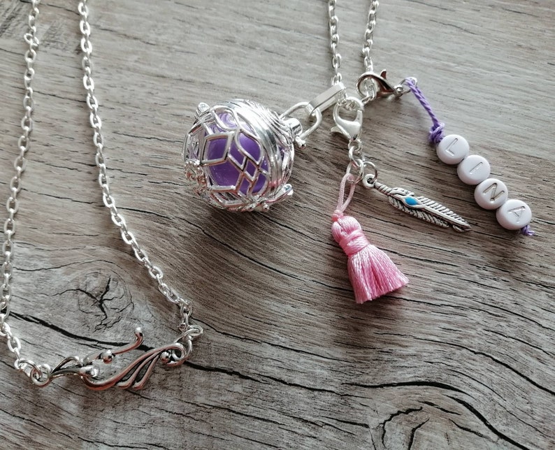 Puede incluir: Un collar de cadena plateada con un colgante de jaula que contiene un orbe de vidrio morado. El collar tambi&eacute;n tiene un borla rosa, un amuleto de pluma plateada y un amuleto con el nombre "LINA" escrito en cuentas blancas.