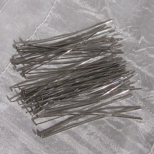 Op de afbeelding: Een verzameling zilverkleurige, rechte metalen pinnen, waarschijnlijk voor knutselen of het maken van sieraden, ligt verspreid op een witte, getextureerde stof. De pinnen zijn dun en lijken van glanzend metaal te zijn, dat licht reflecteert.