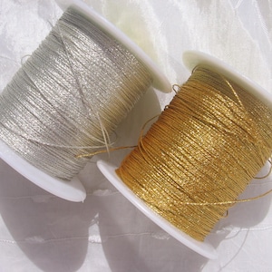 Könnte beinhalten: Zwei Spulen mit Metallic-Garn, eine in Silber und eine in Gold, sind auf weiße Kunststoffspulen gewickelt. Das Garn hat eine reflektierende, schimmernde Textur und ist fest um jede Spule gewickelt, wobei lose Fäden an der Oberfläche sichtbar sind.