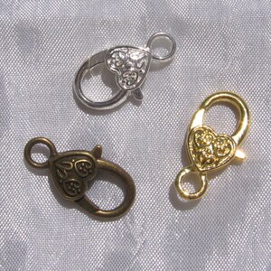 Puede incluir: Tres cierres en forma de corazón en plata, oro y bronce. Cada cierre tiene un diseño de corazón decorativo y un cierre de gancho.