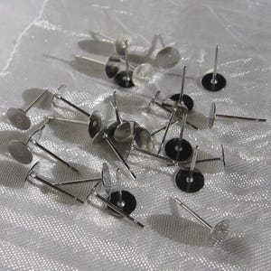 May include: A scattering of silver earring posts, each with a flat, circular back and a slender, straight post. The posts are arranged on a white, textured surface, creating a bright, reflective display. These components are suitable for DIY jewellery making.