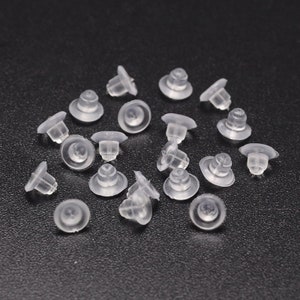 May include: A close-up of a pile of clear plastic earring backs. The backs are small and round, with a small post in the centre.