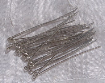 May include: A pile of silver-colored, thin, straight, metal pins with a loop at one end. The pins are arranged in a random pattern on a white fabric surface.