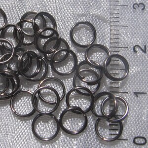 May include: A close-up of a pile of black jump rings. The rings are about 1/2 inch in diameter and are arranged in a random pattern. A ruler is visible in the background, with the numbers 0, 1, 2, and 3 visible.