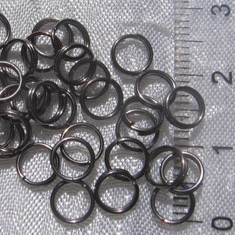 May include: A close-up of a pile of black jump rings. The rings are about 1/2 inch in diameter and are arranged in a random pattern. A ruler is visible in the background, with the numbers 0, 1, 2, and 3 visible.