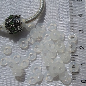 May include: A collection of translucent, donut-shaped beads scattered on a white surface. A silver charm with green accents and a silver bracelet are visible in the background. The beads appear to be made of a soft, opalescent material.