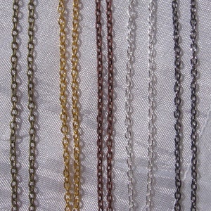 Peut inclure: Cinq chaînes de différentes couleurs, dont le bronze, l'or, le cuivre, l'argent et le noir, sont présentées sur un fond de tissu blanc.