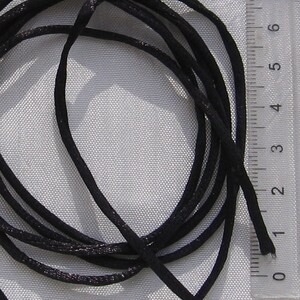 Puede incluir: Primer plano de un cord&oacute;n de sat&eacute;n negro, que mide aproximadamente 2,5 cm de largo, sobre un fondo de tela blanca.