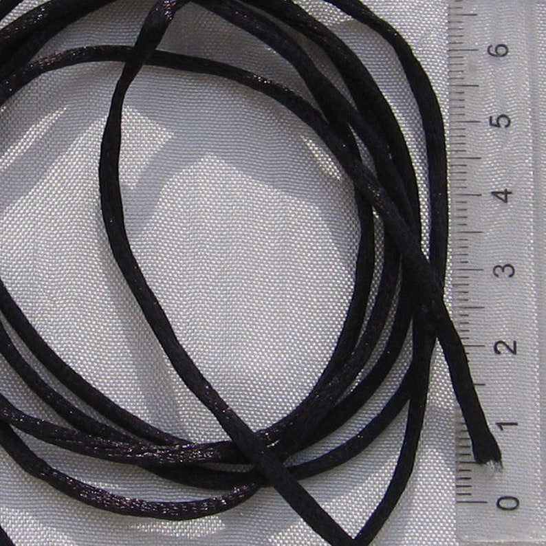 Puede incluir: Primer plano de un cord&oacute;n de sat&eacute;n negro, que mide aproximadamente 2,5 cm de largo, sobre un fondo de tela blanca.