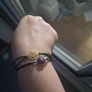 Pu&ograve; includere: Due braccialetti su un polso. Un braccialetto ha un ciondolo in filigrana dorata e l'altro ha un ciondolo in pietra preziosa viola. Entrambi i braccialetti hanno cordini neri.