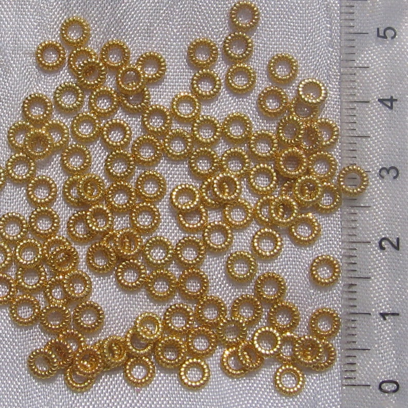 May include: A large quantity of small, gold-colored jump rings. The rings are scattered on a white fabric surface. A ruler is visible in the background.