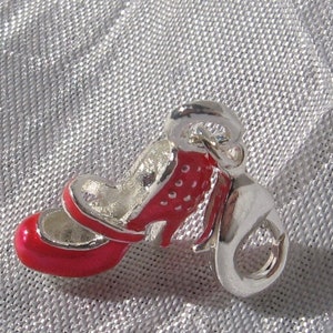 K&ouml;nnte beinhalten: Ein silberfarbener Metallanh&auml;nger in Form eines hochhackigen Schuhs mit leuchtend roter Emaille-Beschichtung an der Au&szlig;enseite. Der Schuh hat eine abgerundete Spitze, einen d&uuml;nnen Riemen &uuml;ber dem Spann und einen kleinen Absatz. Ein Karabiner ist befestigt.
