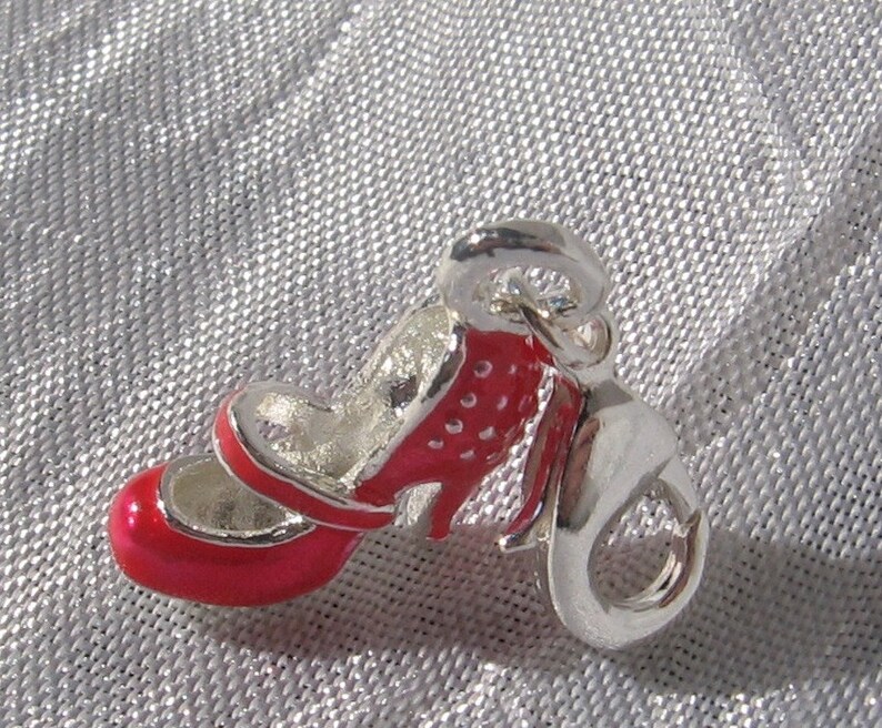 K&ouml;nnte beinhalten: Ein silberfarbener Metallanh&auml;nger in Form eines hochhackigen Schuhs mit leuchtend roter Emaille-Beschichtung an der Au&szlig;enseite. Der Schuh hat eine abgerundete Spitze, einen d&uuml;nnen Riemen &uuml;ber dem Spann und einen kleinen Absatz. Ein Karabiner ist befestigt.