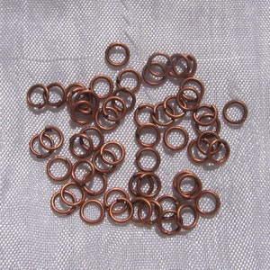 Puede incluir: Un conjunto de pequeños anillos de salto de color cobre dispersos sobre una superficie gris claro. Los anillos son circulares con una pequeña abertura, lo que sugiere su uso en la fabricación de joyas o manualidades. El cálido tono metálico contrasta con el fondo neutro.